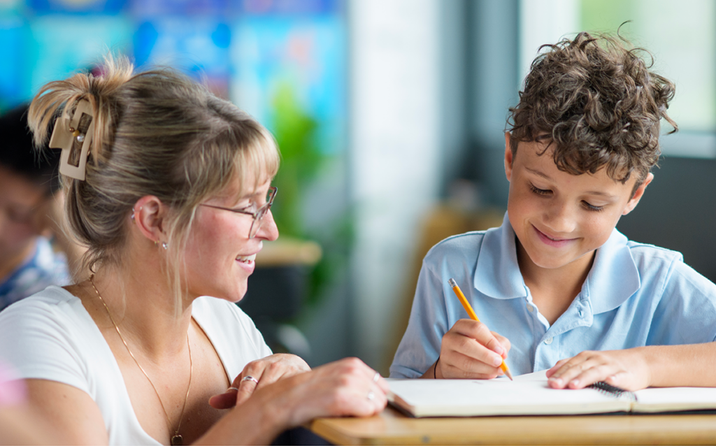 A teacher is assisting an elementary student who is writing in a notebook at a classroom desk.