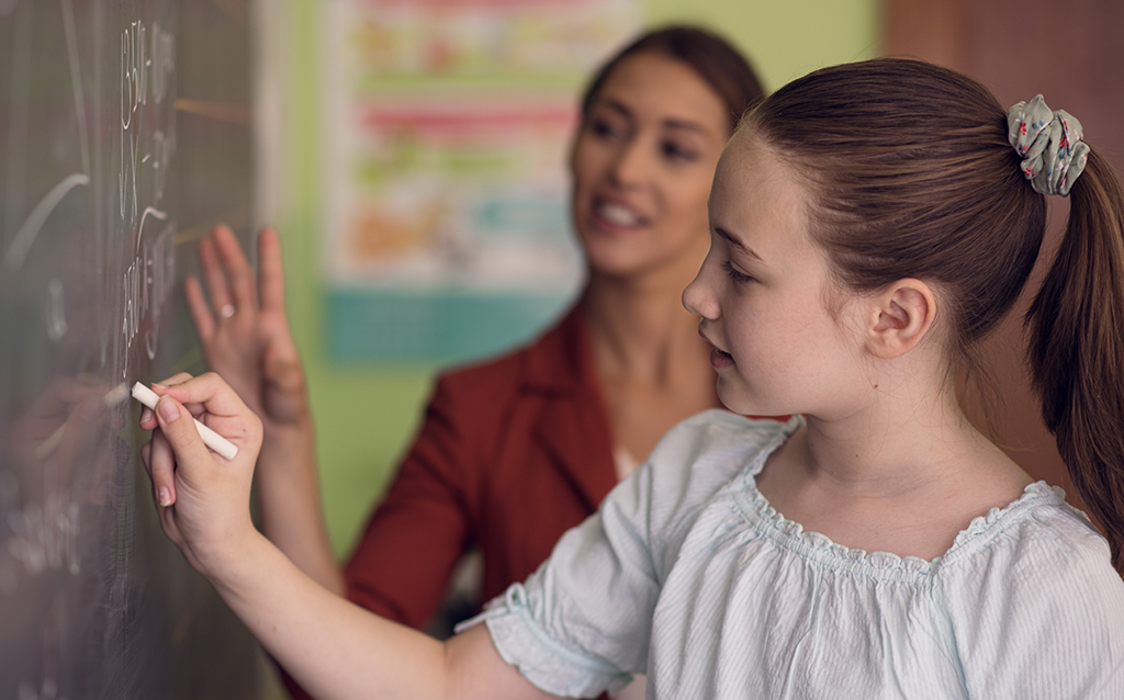 Student writing on a classroom chalkboard while a teacher provides guidance.